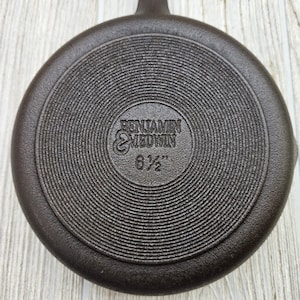 May include: A dark brown cast iron pan with a handle. The bottom of the pan has concentric circular ridges and the text "BENJAMIN & MEDWIN 6 1/2"" is embossed in the center.