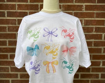 Peut inclure: T-shirt blanc avec un motif graphique coloré d'abeilles et de nœuds. Les abeilles sont de différentes couleurs, notamment jaune, vert, violet et rouge. Les nœuds sont dans des tons de violet, bleu et jaune.