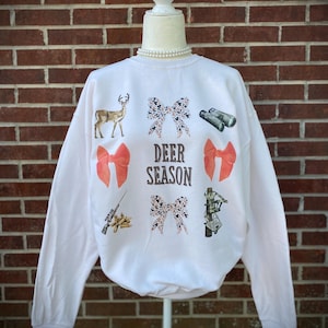 May include: White sweatshirt with a "Deer Season" graphic. The design includes a deer, bows, binoculars, a rifle, and a hunter on an ATV. The bows have a leopard print pattern. The sweatshirt is displayed on a mannequin.