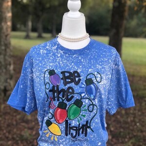 Peut inclure: Un t-shirt bleu avec un motif de peinture blanche. Le t-shirt a un graphique d'ampoules color&eacute;es avec le texte "Soyez la lumi&egrave;re" et "Matthieu 5:14".