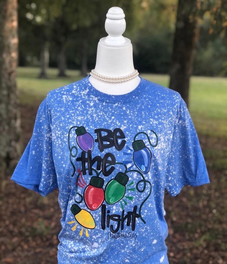 Peut inclure: Un t-shirt bleu avec un motif de peinture blanche. Le t-shirt a un graphique d'ampoules color&eacute;es avec le texte "Soyez la lumi&egrave;re" et "Matthieu 5:14".