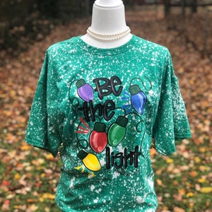Peut inclure: T-shirt vert avec un motif de peinture blanche. Le t-shirt pr&eacute;sente un motif de lampes color&eacute;es avec le texte "Soyez la lumi&egrave;re" et "Matthieu 5:14".