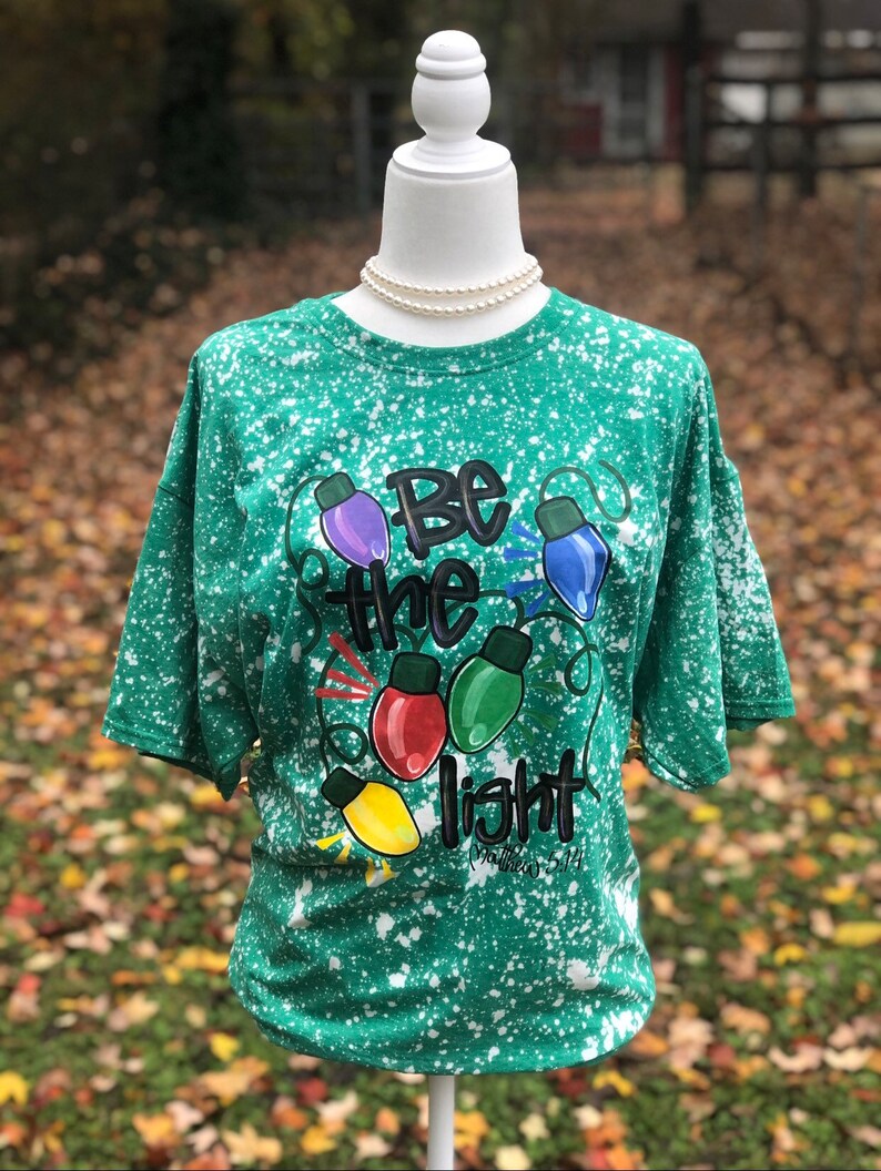 Peut inclure: T-shirt vert avec un motif de peinture blanche. Le t-shirt pr&eacute;sente un motif de lampes color&eacute;es avec le texte "Soyez la lumi&egrave;re" et "Matthieu 5:14".