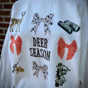 May include: Cream-colored sweatshirt with deer-themed graphics. Features a deer, bows, binoculars, a rifle, and a crossbow. The words "DEER SEASON" are printed in the center. A pearl necklace is visible at the neckline.