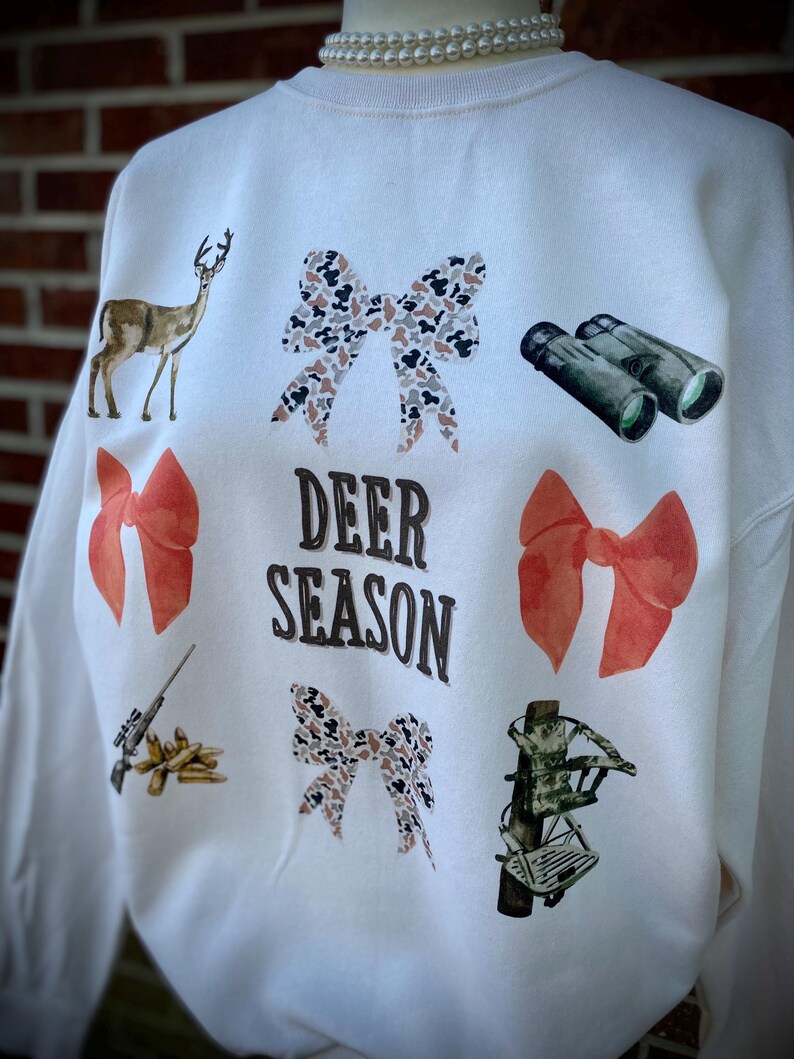 May include: Cream-colored sweatshirt with deer-themed graphics. Features a deer, bows, binoculars, a rifle, and a crossbow. The words "DEER SEASON" are printed in the center. A pearl necklace is visible at the neckline.