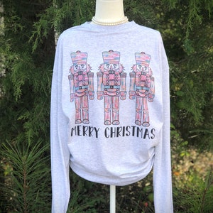 May include: A gray long-sleeved shirt with a graphic of three colorful nutcracker figures and the text "Merry Christmas".