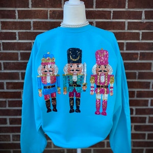 May include: A turquoise blue sweatshirt with three sequined nutcracker figures on the front. The nutcrackers are wearing blue, green, and pink outfits.