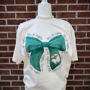 May include: A cream-colored t-shirt with a green bow and the text "Let's go eagles" repeated around the bow. An eagle mascot is in the center of the bow.