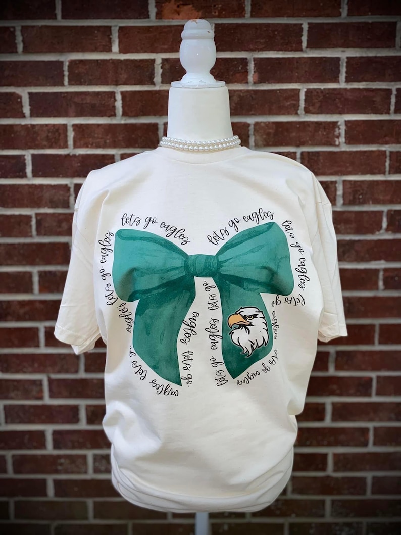 May include: A cream-colored t-shirt with a green bow and the text "Let's go eagles" repeated around the bow. An eagle mascot is in the center of the bow.