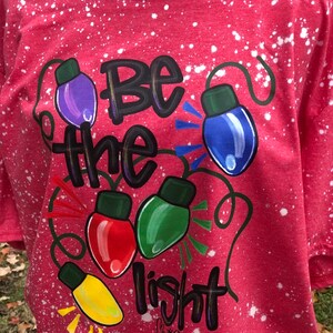 Peut inclure: T-shirt rouge avec un motif de peinture blanche. Le t-shirt a un motif de lampes color&eacute;es avec les mots "Soyez la lumi&egrave;re" et l'&Eacute;criture "Matthieu 5:14".