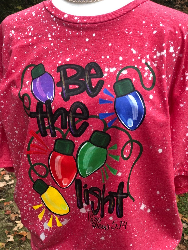 Peut inclure: T-shirt rouge avec un motif de peinture blanche. Le t-shirt a un motif de lampes color&eacute;es avec les mots "Soyez la lumi&egrave;re" et l'&Eacute;criture "Matthieu 5:14".