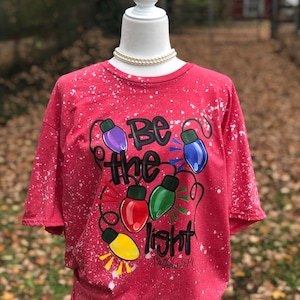 Peut inclure: T-shirt rouge avec un motif &eacute;clabouss&eacute; de javel blanche. Le t-shirt pr&eacute;sente une guirlande de lampes color&eacute;es avec les mots "Soyez la lumi&egrave;re" et l'&Eacute;criture "Matthieu 5:14".