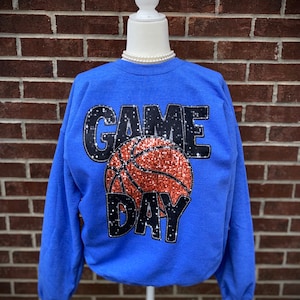 May include: A royal blue sweatshirt with the words "GAME DAY" in black glitter with a basketball in the middle. The basketball is orange and black glitter.