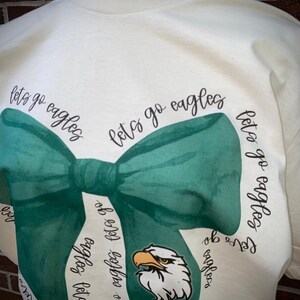 May include: A white t-shirt with a green bow tie design and the text "let's go eagles" repeated around the bow tie. The bow tie has a green eagle head in the center.