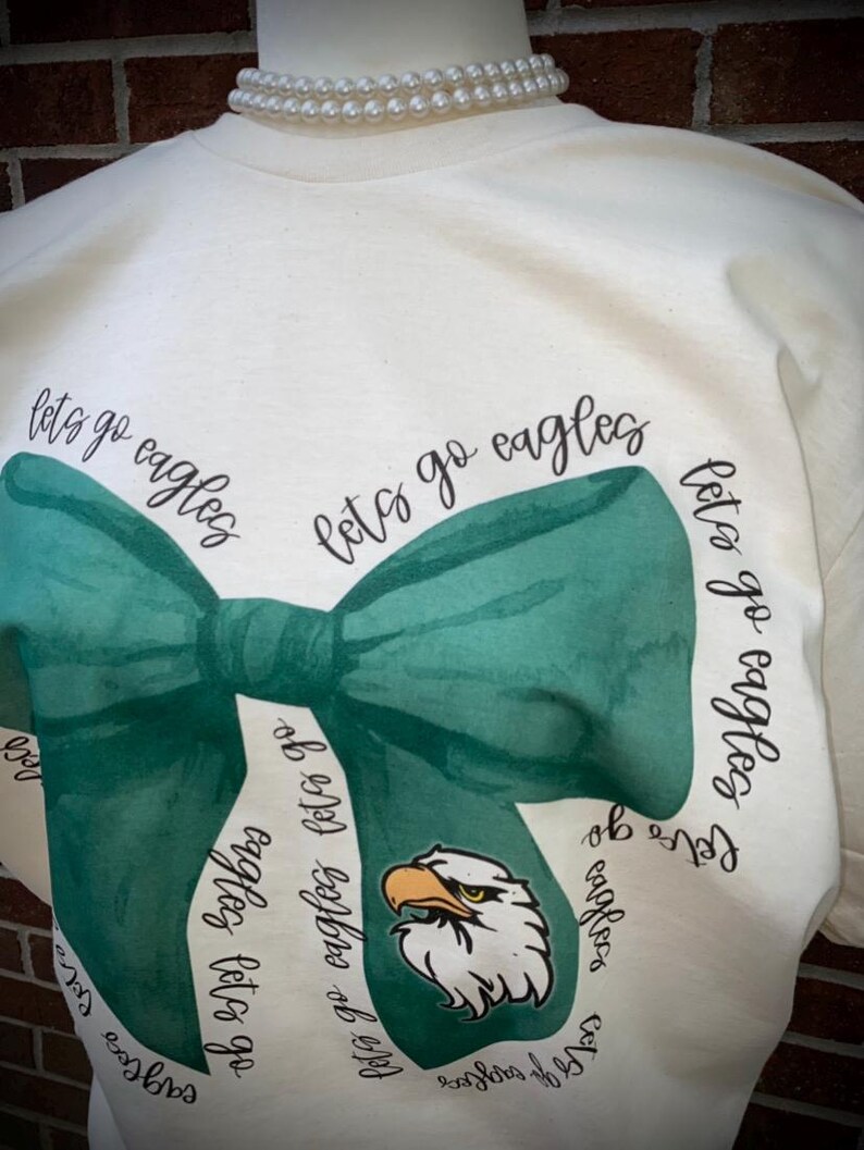 May include: A white t-shirt with a green bow tie design and the text "let's go eagles" repeated around the bow tie. The bow tie has a green eagle head in the center.