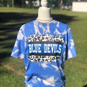 May include: Blue tie-dye t-shirt with a white and black leopard print design and the text "BLUE DEVILS" in blue glitter.