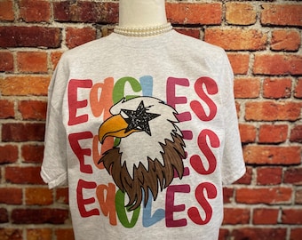 Peut inclure: Un t-shirt gris clair avec le mot "EAGLES" en lettres colorées. Le t-shirt présente un graphisme d'un aigle avec une étoile sur la tête. L'aigle a un bec jaune et des plumes marron. Le t-shirt est présenté sur un mannequin.