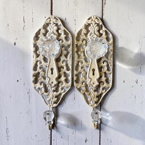 May include: Two matching wall hooks featuring an off-white, ornate metal backplate with a keyhole design. Each hook is accented with a clear, faceted glass knob at the top and a smaller, clear glass hook at the bottom for hanging items.