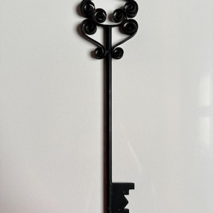 May include: Black metal decorative key with a scrolled top and a rectangular key bit. The key's design features a long, straight stem and decorative swirls at the top. The key is set against a white background.