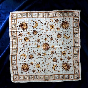 Puede incluir: Un pañuelo cuadrado blanco con un estampado celestial de soles, lunas, estrellas y planetas dorados. El borde contiene un patrón repetitivo de símbolos del zodiaco en cuadrados pequeños. El texto GIANNI VERSACE aparece en la esquina inferior.