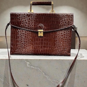 May include: A brown briefcase with a crocodile skin texture. The bag has a gold-colored handle and a long, thin shoulder strap. The briefcase has a rectangular shape and a clasp closure.