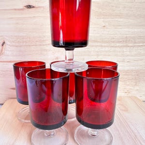May include: A set of six vintage red wine glasses. The glasses have a clear stem and base, with a deep red bowl. The glasses are arranged on a light wooden surface, with a natural wood grain background.