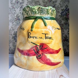 May include: A ceramic jar in the shape of a sack, painted yellow with red chili peppers. The jar has the words "Pomme de Terre" and "Pays Basque" written on it. The top is green and the jar is sitting on a wooden surface.