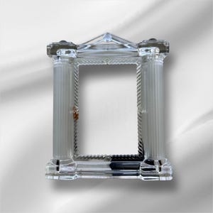May include: A clear glass picture frame with a classical column design. The frame has two fluted columns, a triangular top, and a decorative rope-like border. The frame is designed to hold a rectangular photo.