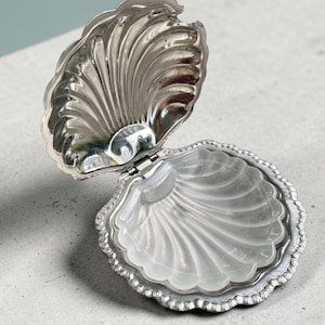 May include: A silver-toned metal clam shell trinket box with a hinged lid. The interior of the bottom half features a translucent, frosted glass insert shaped like a shell. The exterior metal surface has a polished, reflective finish with ridges.