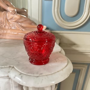 May include: A vibrant red glass jar with a lid, featuring intricate embossed patterns. The jar has a round shape with a small knob on top of the lid. The jar is sitting on a white marble surface.