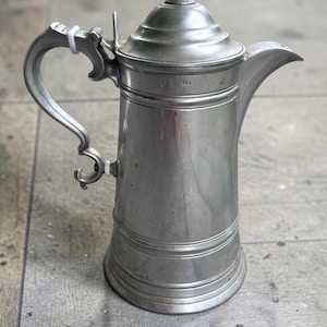 May include: A silver-toned metal pitcher with a tall cylindrical body, a flared base, and a curved handle. The pitcher features a hinged lid with a pointed finial and a spout. A small stamp is visible on the upper body near the handle attachment.