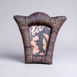 May include: A trapezoidal picture frame constructed from dark brown woven rattan or wicker material. The frame features a flared top edge and contains a central insert displaying a dark gray background with a light tan, abstract organic pattern.