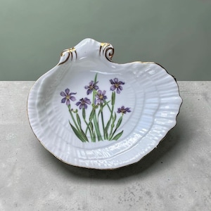 May include: A white, shell-shaped dish with a gold rim and a floral design. The dish features a cluster of purple iris flowers with green stems and leaves. The dish is made of porcelain.