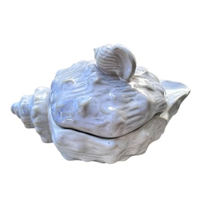 May include: A light blue ceramic seashell-shaped container with a lid. The container has a textured surface and a small seashell detail on top of the lid. The container is on a white background.