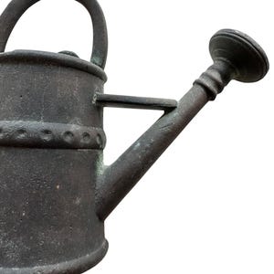 May include: A weathered, dark gray metal watering can with a curved handle and a spout. The can has a decorative band around the middle and a small circular base. The spout has a round, ridged head.