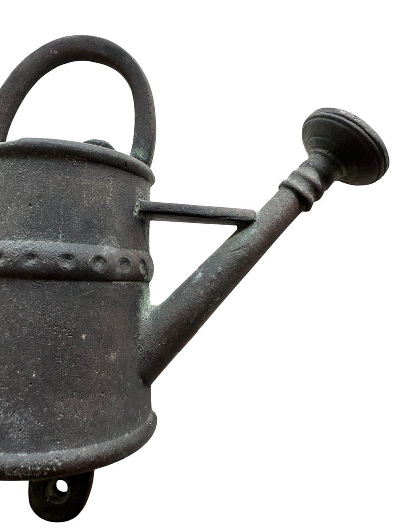 May include: A weathered, dark gray metal watering can with a curved handle and a spout. The can has a decorative band around the middle and a small circular base. The spout has a round, ridged head.