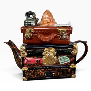 May include: A ceramic teapot shaped like a stack of two vintage suitcases. The bottom suitcase is black and brown with gold latches and labels reading First Class GB and London & SW Railway to Portsmouth. The top suitcase holds a kettle and hat.