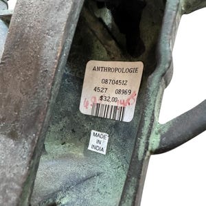 May include: A close-up of a weathered, bronze-colored metal object with a textured surface. The object has a handle and a rectangular shape. A label from Anthropologie is visible, along with a "Made in India" sticker.
