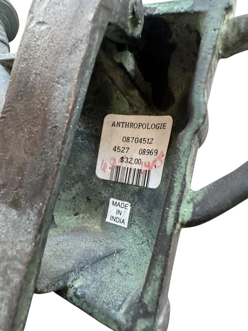May include: A close-up of a weathered, bronze-colored metal object with a textured surface. The object has a handle and a rectangular shape. A label from Anthropologie is visible, along with a "Made in India" sticker.