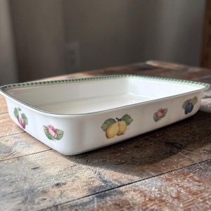 May include: Rectangular white ceramic baking dish with fruit illustrations. The dish features a green border and is decorated with images of plums, lemons, and other fruits. The dish is placed on a wooden surface.