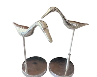 Puede incluir: Dos esculturas de pájaros plateados. Cada pájaro tiene un cuello largo y curvado y está montado sobre una fina varilla de metal unida a una base circular. Los pájaros miran en diferentes direcciones, creando una exhibición dinámica.