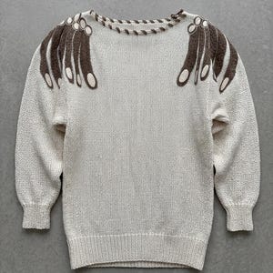 Puede incluir: Suéter color crema con bordados marrones en forma de manos en los hombros. El suéter tiene cuello redondo y mangas largas con puños y dobladillo acanalados. Los diseños de manos tienen detalles ovalados blancos.