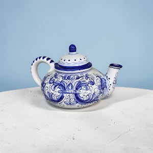 May include: A white ceramic teapot featuring intricate blue hand-painted floral and scrollwork patterns. The teapot has a rounded body, a curved spout, a sturdy handle with blue stripes, and a matching lid with a small knob on top.
