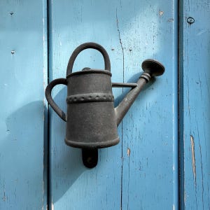 May include: A dark gray, cast iron watering can shaped door knocker. The door knocker is mounted on a weathered, light blue wooden door. The watering can has a handle, spout, and decorative band.