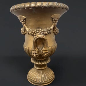Puede incluir: Una urna de color dorado con un borde ancho y estriado y una base de pedestal. El cuerpo presenta relieves de guirnaldas drapeadas y dos asas, cada una adornada con una cara esculpida en la base. La superficie tiene un acabado metálico.