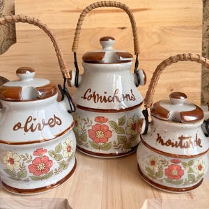 Puede incluir: Tres tarros de cerámica para condimentos con tapas y asas tejidas. Los tarros son blancos con detalles marrones y estampados florales. Los tarros están etiquetados como "olivas", "cornichons" y "mostaza".