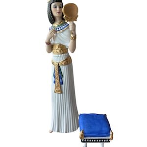 Puede incluir: Una figura de porcelana de Cleopatra sosteniendo un espejo dorado, vestida con un vestido blanco con detalles dorados y azules. Hay un pequeño taburete azul y dorado a su lado. La figura tiene cabello negro y una corona dorada.