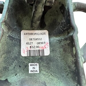 May include: Close-up of a weathered, green-toned metal object with a label from Anthropologie. The label includes a barcode and the text "MADE IN INDIA". The object has a textured surface and appears to be a decorative item.