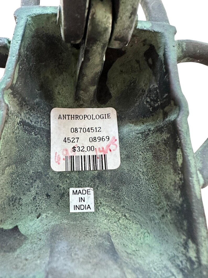 May include: Close-up of a weathered, green-toned metal object with a label from Anthropologie. The label includes a barcode and the text "MADE IN INDIA". The object has a textured surface and appears to be a decorative item.