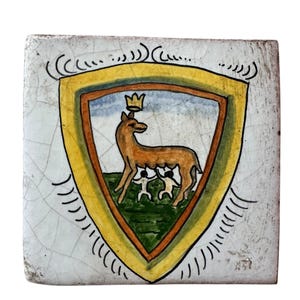 May include: Square ceramic tile with a hand-painted coat of arms. The shield features a brown deer wearing a crown, with two figures below, set against a green and blue background, framed by a yellow border. The tile has a cracked, aged appearance.